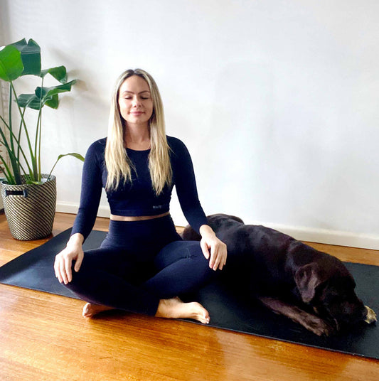 Woman practicing meditation at home in black outfit with the good company of a chocolate labrador dog, showing her confidence in white artica women's sportswear